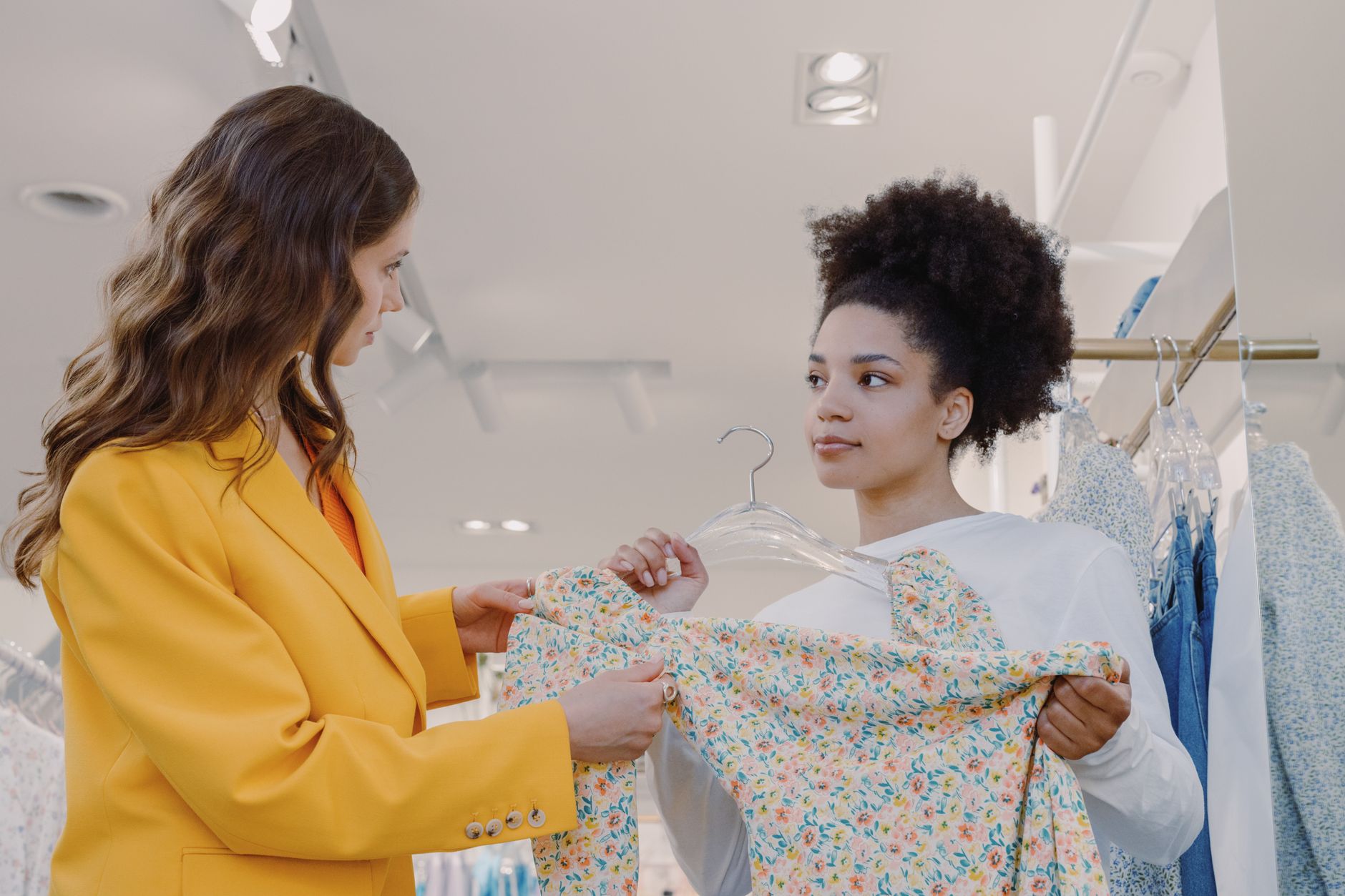 Two women in a bright modern clothing shop, one in a yellow blazer holding up a floral dress on a hanger for the other to look at