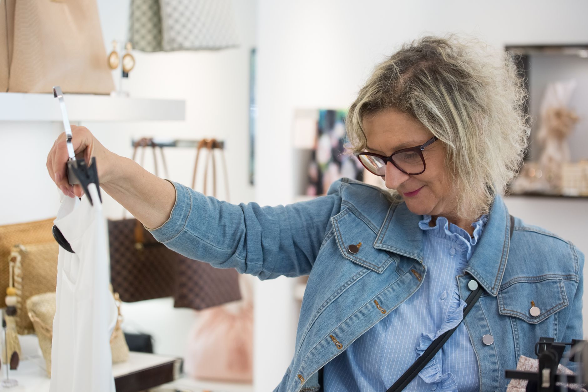 A woman in a denim jacket and glasses lifting a white blouse on a hanger from a rail in a bright boutique, with handbags displayed on shelves behind her
