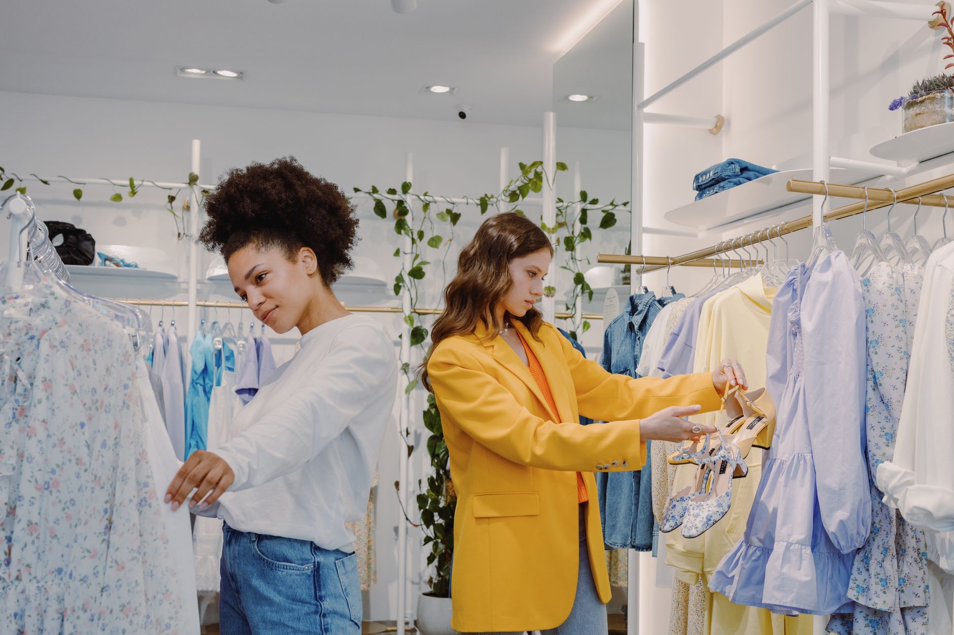 Two women browsing a rail of pastel dresses in a bright, plant-filled clothing boutique