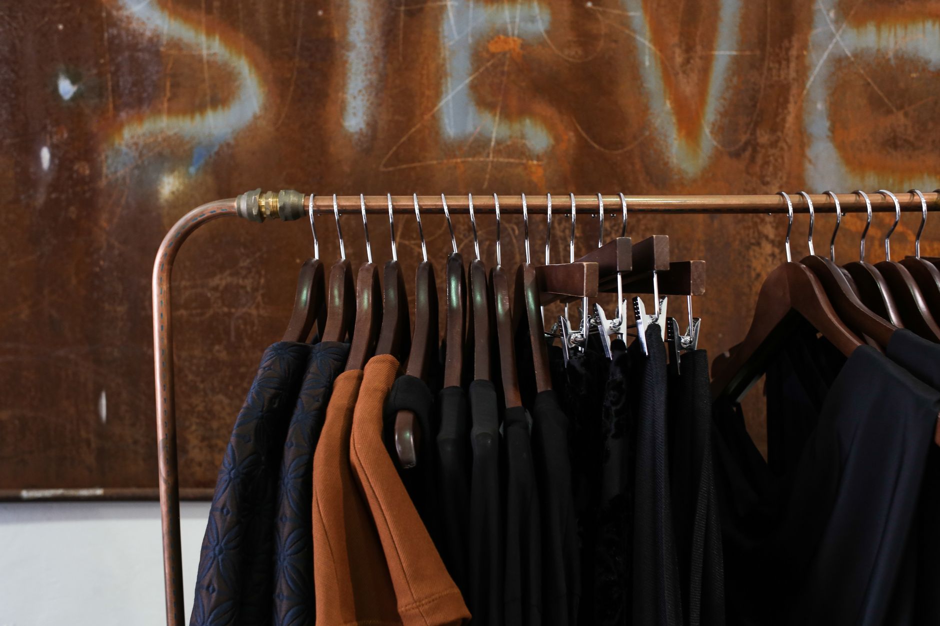 A copper clothing rail hung with dark and rust-coloured garments on wooden hangers, standing against a large rusted steel wall inside a boutique