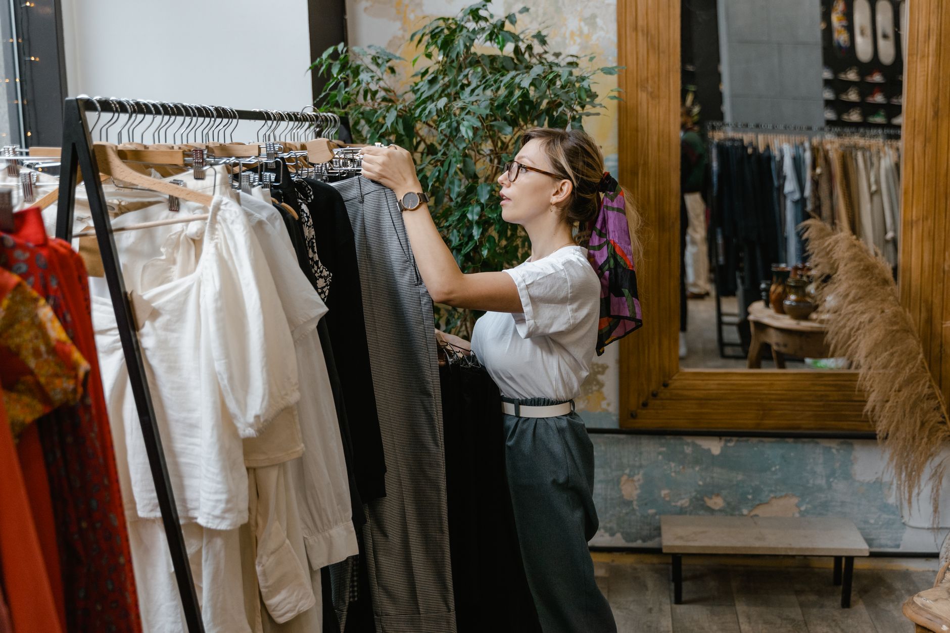 A woman in glasses and a white t-shirt browsing a rail of clothes in a small boutique with a large wooden-framed mirror and a potted tree behind her