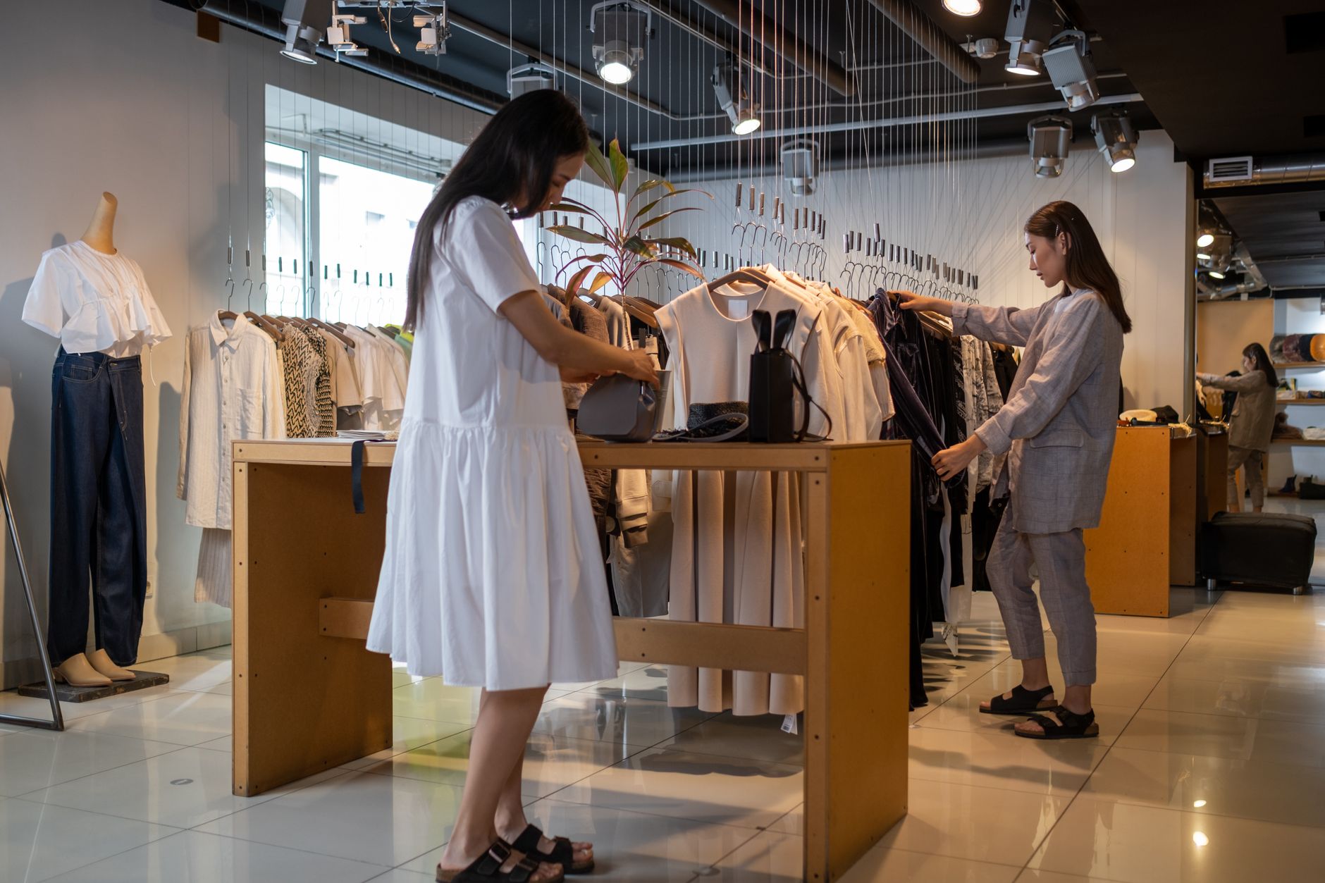 Two shoppers browsing rails of neutral-toned clothing in a bright, minimally furnished store with polished floors