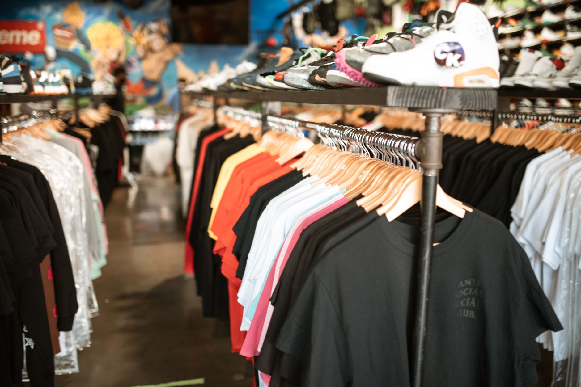 ストリートウェアの店内。木製ハンガーに黒・白・赤・ピンクの無地Tシャツが並び、その上の棚にはスニーカーが置かれ、奥の壁にはアニメ風の壁画が描かれている