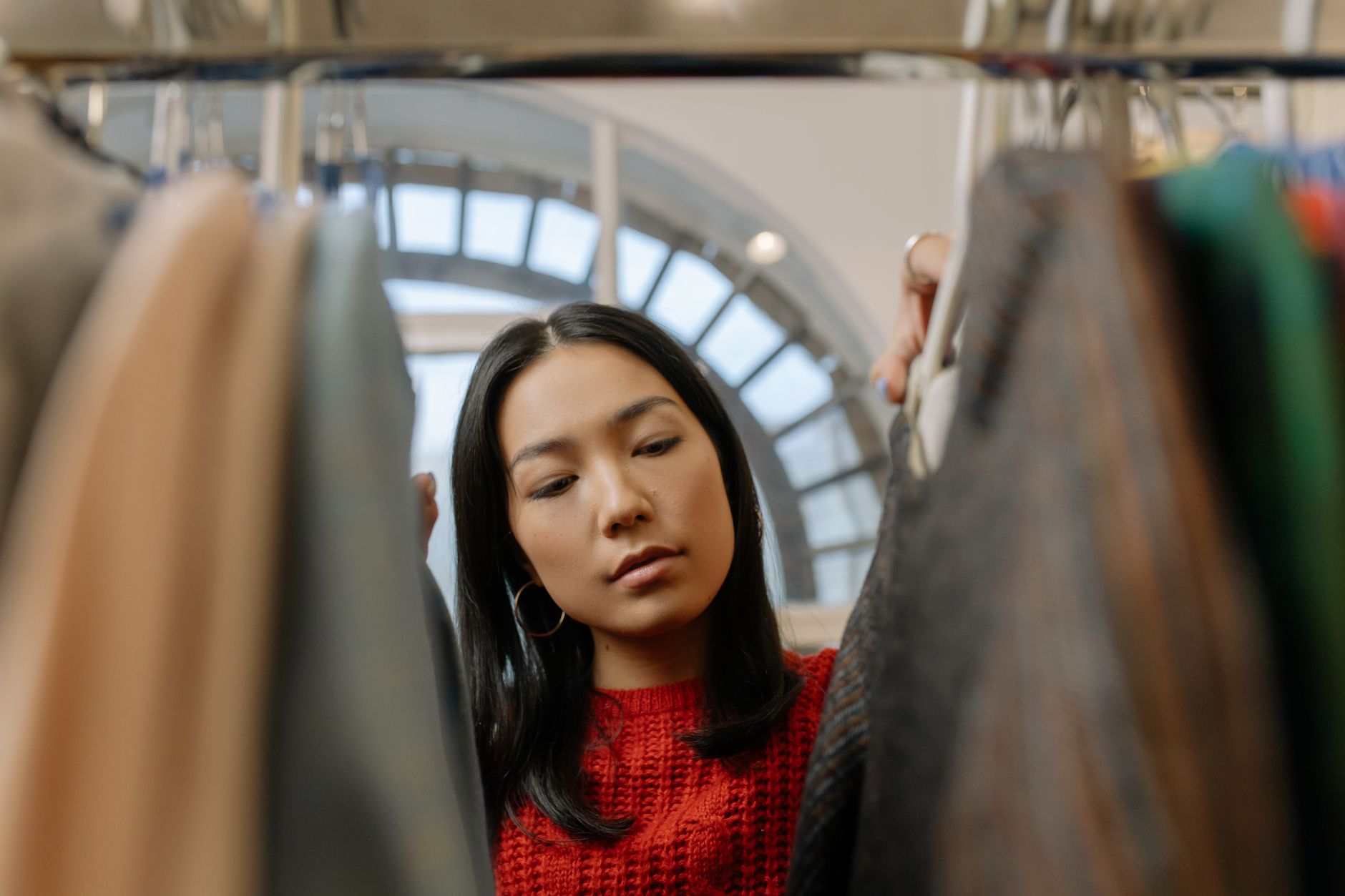 A woman in a red knitted jumper and gold hoop earrings looking through coats on a clothing rail, under an arched glass roof window