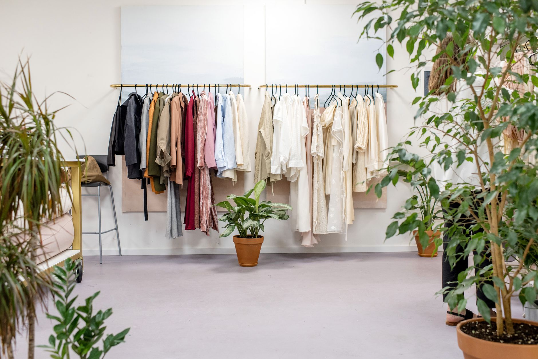 A bright boutique interior with two brass rails of coats, shirts and dresses in muted colours against a white wall, potted plants and a small stool in the foreground
