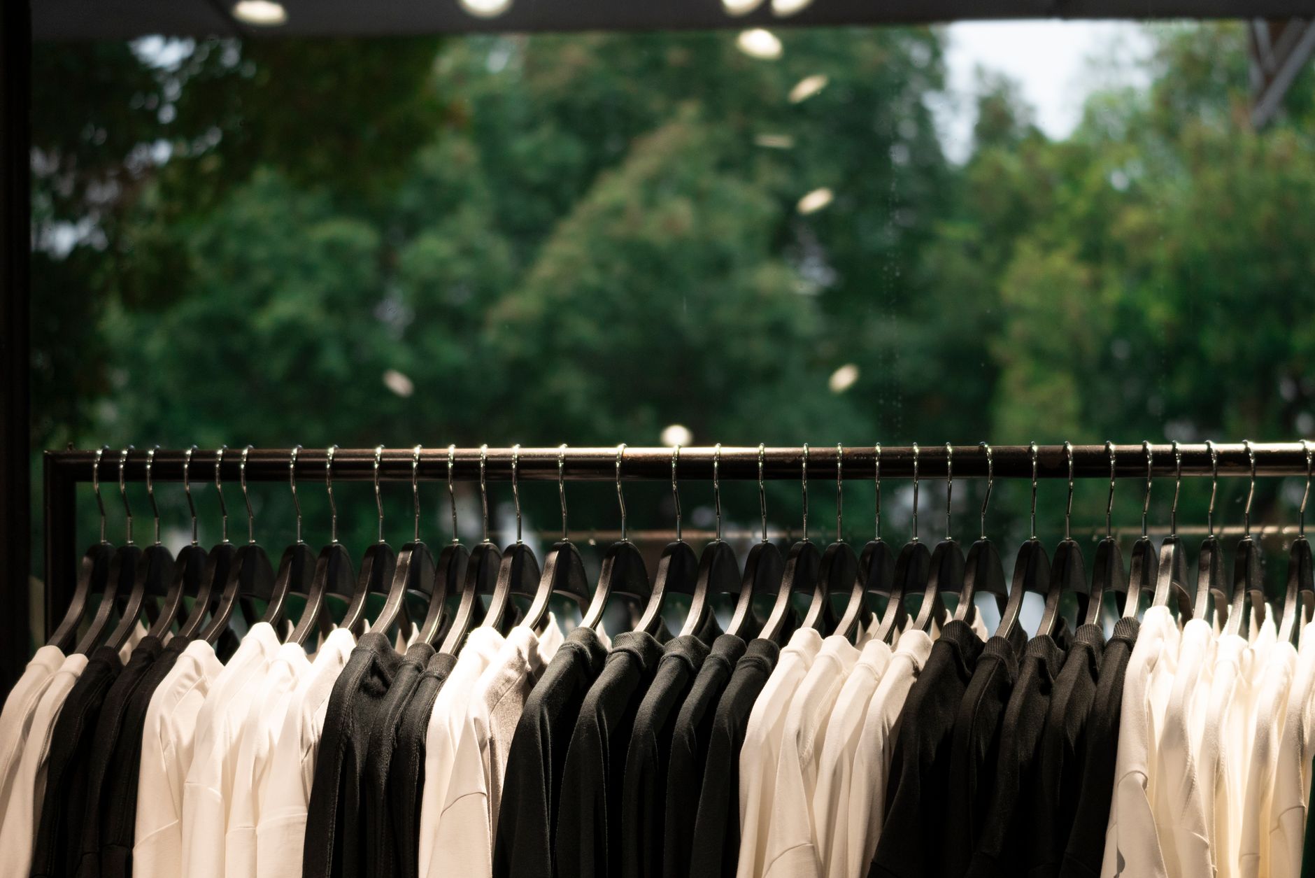 A boutique rail seen from inside the shop: a long steel bar of identical dark hangers carrying alternating blocks of cream and black garments, with a floor-to-ceiling window behind it opening onto sunlit green trees