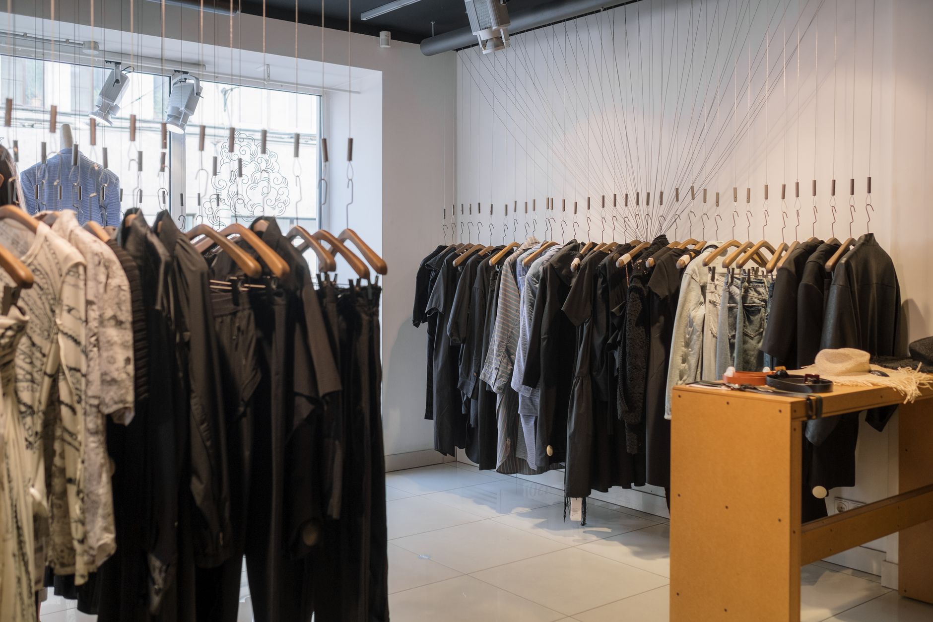 A bright minimalist boutique: rails of mostly black and grey clothes on wooden hangers, a wall hung with dozens of empty wire hangers on long cords, track spotlights above a pale tiled floor, and a mustard-yellow table holding a straw hat, belts and sunglasses
