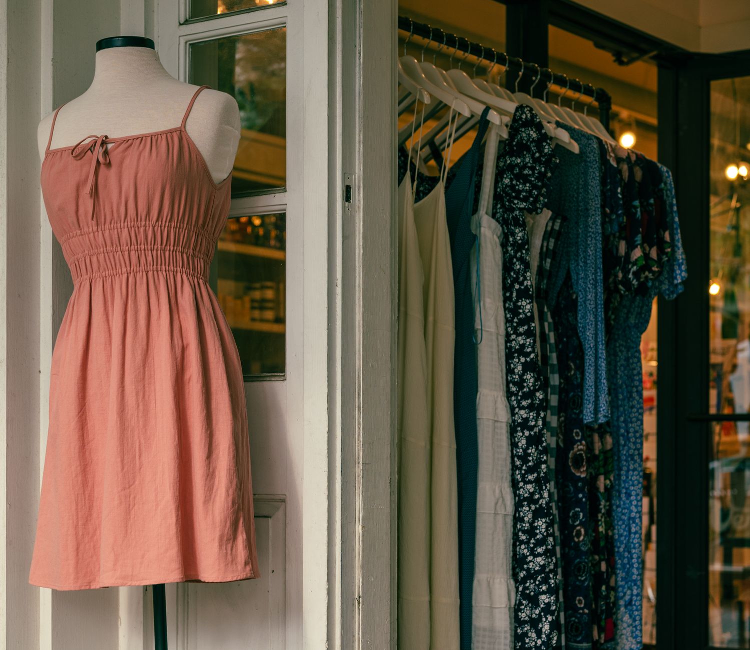 A dusty-pink sleeveless dress on a headless mannequin outside a shop with white-painted glazed doors, beside a rail of plain and floral dresses on wooden hangers under warm interior lights