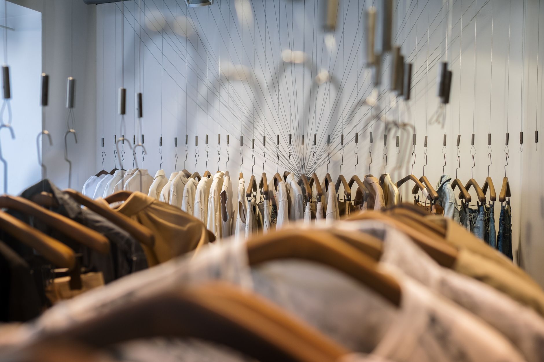 The inside of a small clothing boutique, rows of shirts and jeans on wooden hangers suspended from thin cables along a white wall, lit softly from above