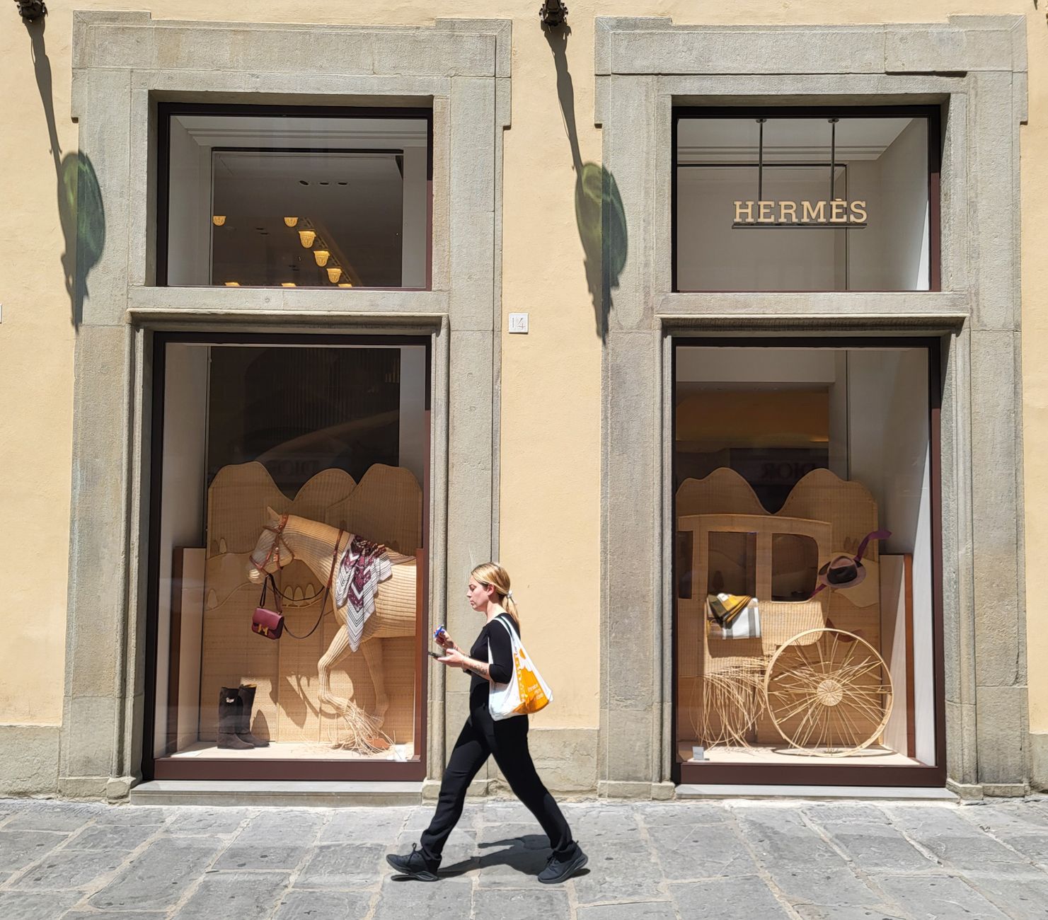 A woman walking past the lit window displays of Hermès and Dior on a sunlit Mediterranean stone street