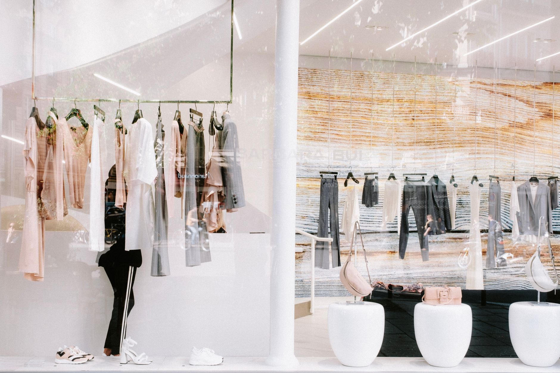 Looking through the window of a bright fashion boutique: pale dresses and grey tailoring on brass rails, handbags on rounded white plinths, and the street reflected across the glass