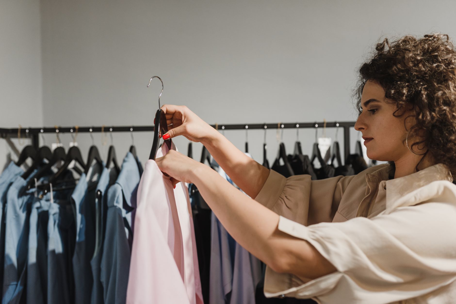 A woman in a cream blouse lifting a pale pink garment on a hanger from a rail of blue and grey shirts in a small, plainly lit clothing shop