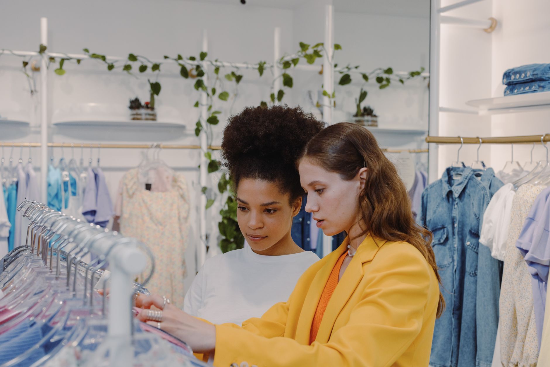 Two women looking through a rail of pastel clothes in a bright white boutique with trailing plants above the rails