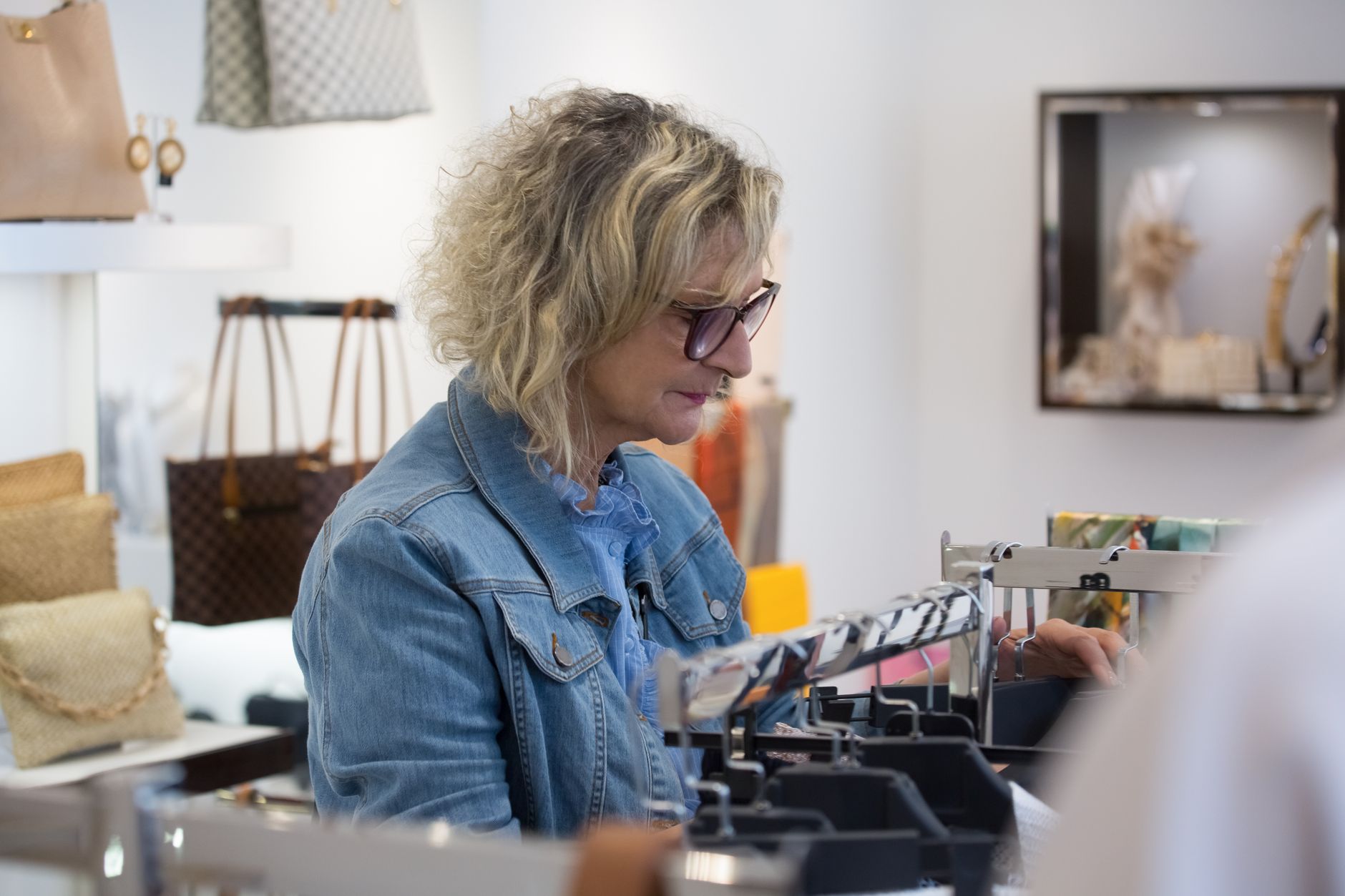 A woman in a denim jacket and glasses looking down at a rail of hangers in a bright boutique, with leather handbags on display shelves behind her