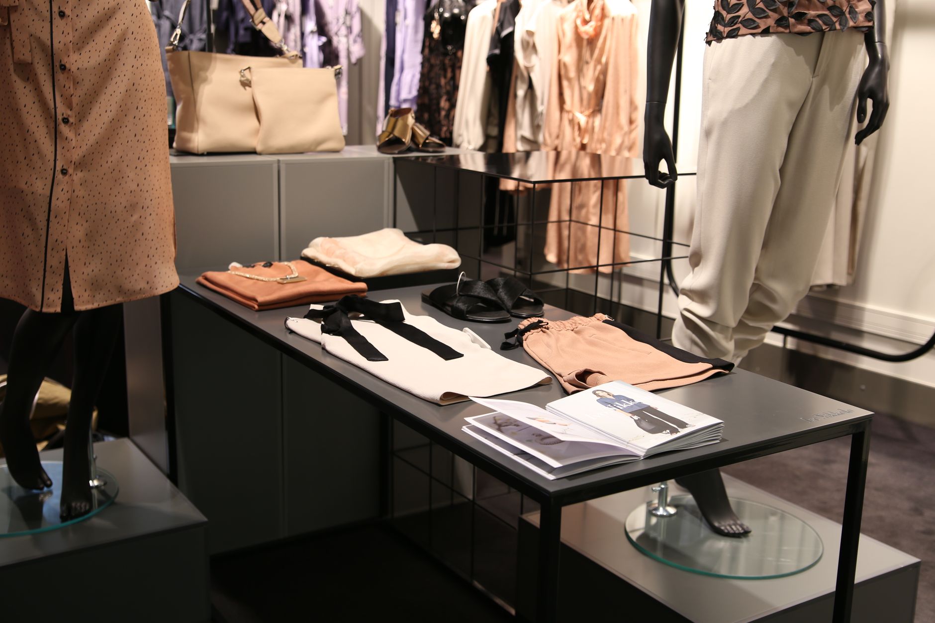 The interior of a fashion boutique, with folded knitwear in blush and cream laid out on a low black display table beside leather sandals and an open lookbook, and mannequins in tailored neutral clothing behind