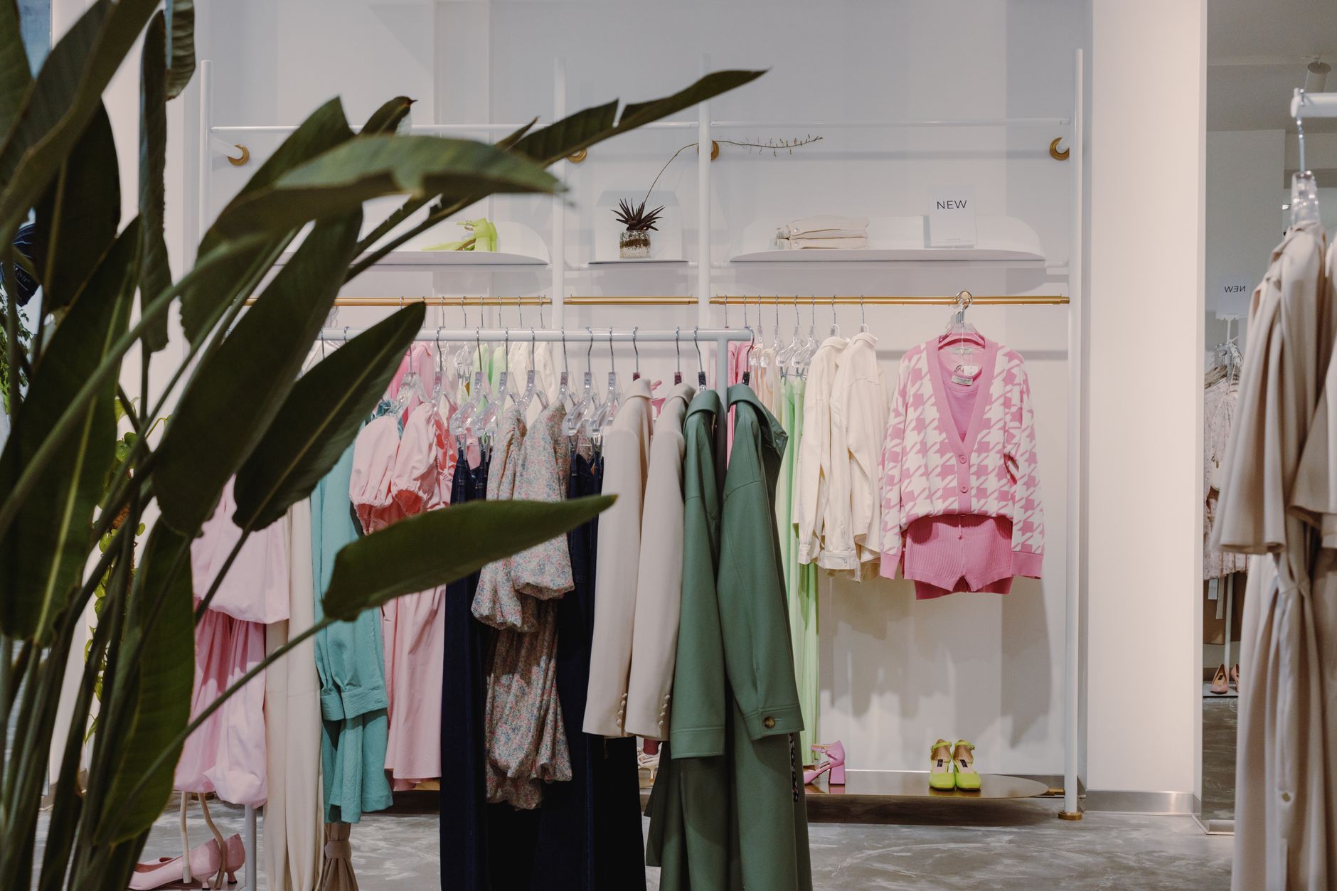 A bright, minimally furnished clothing boutique with a rail of pastel pink, mint and cream garments under a brass rail, a houndstooth cardigan on display and large green plant leaves in the foreground