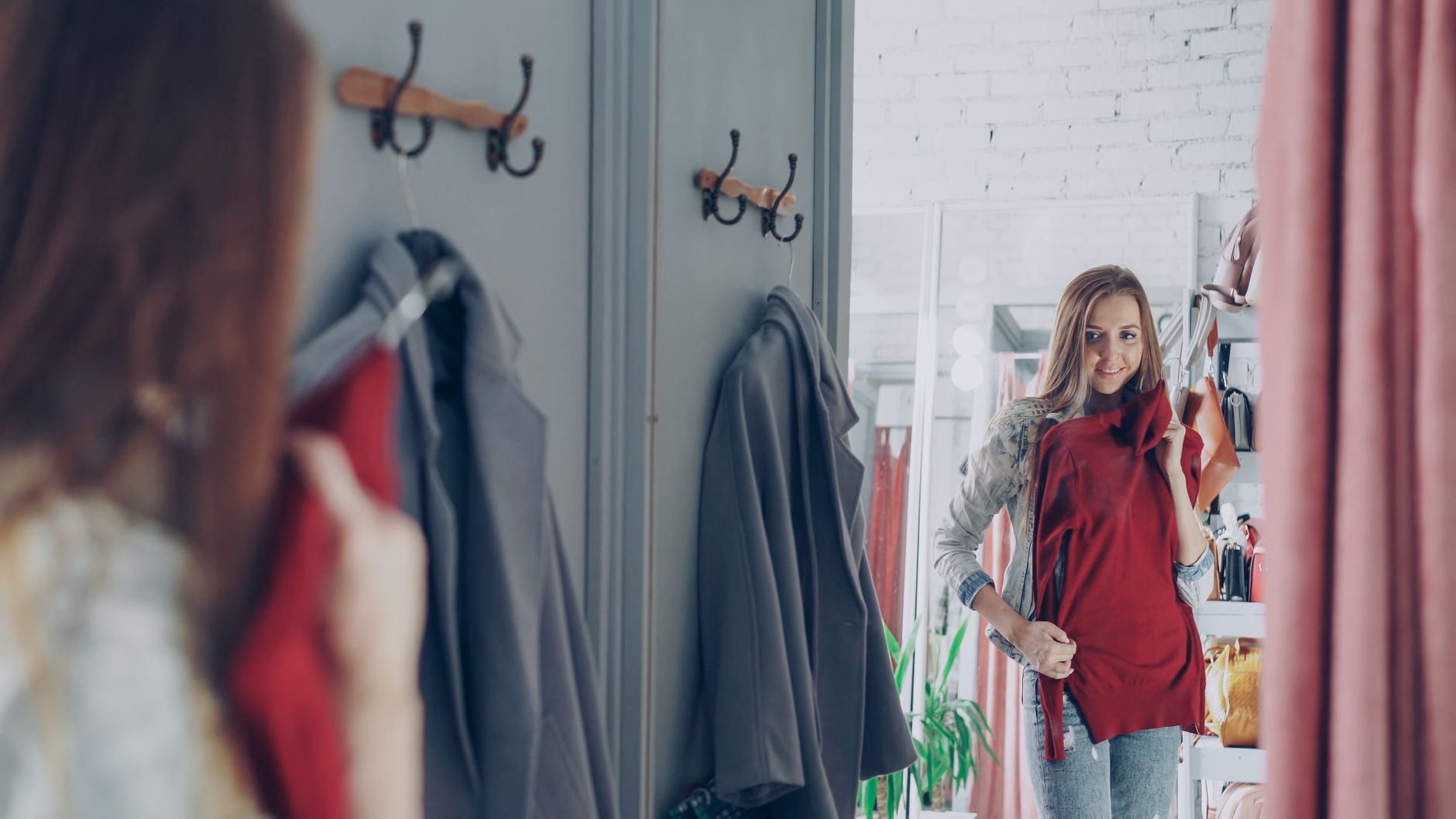 A woman holding a red top against herself in front of a fitting-room mirror in a bright boutique with clothes on rails behind her