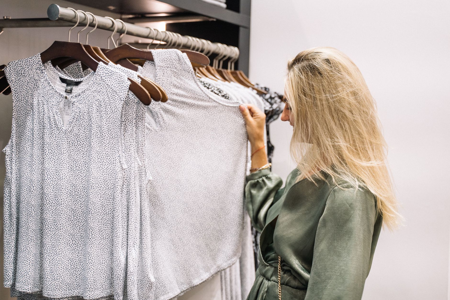 A woman in a green satin dress looking through a rail of white patterned sleeveless tops on wooden hangers in a bright, minimal boutique