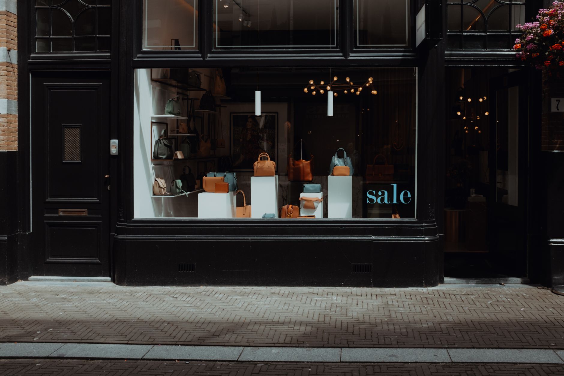 A dark-painted boutique storefront with tan, olive and pale blue leather handbags arranged on white plinths behind the window, and a blue sale decal on the glass